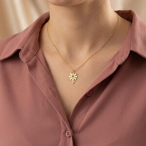 May include: A gold daisy pendant necklace. The necklace is worn against a pink shirt. The daisy pendant is a delicate, floral design. The chain is thin and subtle, complementing the pendant. The necklace is a simple, elegant piece of jewellery.