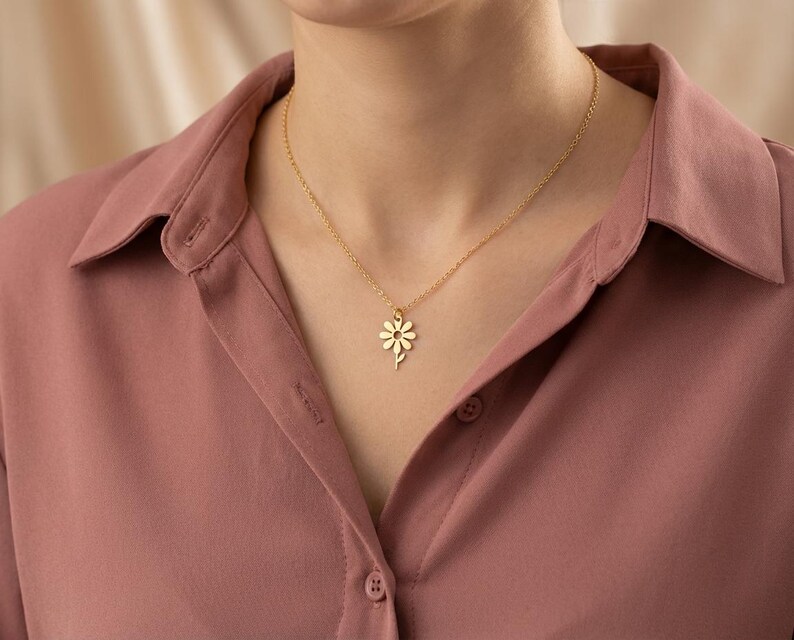 May include: A gold daisy pendant necklace. The necklace is worn against a pink shirt. The daisy pendant is a delicate, floral design. The chain is thin and subtle, complementing the pendant. The necklace is a simple, elegant piece of jewellery.