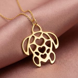 May include: A gold-colored pendant necklace featuring a turtle design with heart-shaped cutouts. The necklace hangs on a delicate chain, and the turtle pendant is the focal point against a pink background. Jewelry.