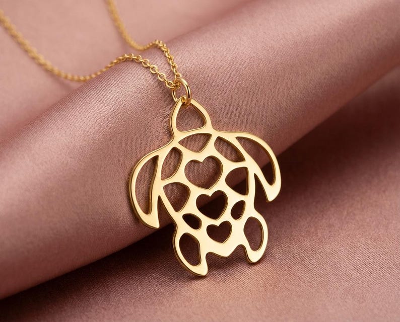 May include: A gold-colored pendant necklace featuring a turtle design with heart-shaped cutouts. The necklace hangs on a delicate chain, and the turtle pendant is the focal point against a pink background. Jewelry.