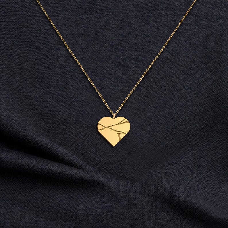 Peut inclure: Un collier avec un pendentif en forme de c&oelig;ur dor&eacute;. Le c&oelig;ur pr&eacute;sente un motif de fissures. La cha&icirc;ne du collier est &eacute;galement dor&eacute;e, pos&eacute;e sur un tissu bleu fonc&eacute;.
