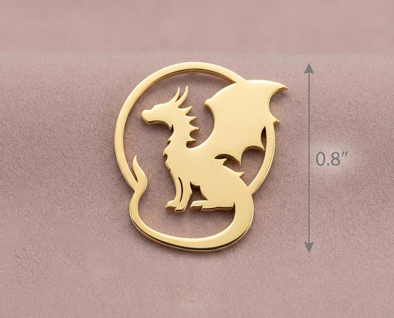 May include: A gold-colored dragon pendant, approximately 0.8 inches in height, is set within a circular frame. The dragon is depicted in a seated pose with detailed wings and a curved tail. The background is a muted pink.
