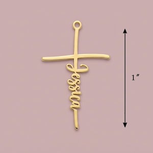 May include: A gold-coloured cross pendant with the name "Jessica" in cursive script. The pendant measures approximately 2.5 cm in length. The background is a muted pink colour.