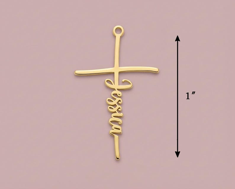 May include: A gold-coloured cross pendant with the name "Jessica" in cursive script. The pendant measures approximately 2.5 cm in length. The background is a muted pink colour.