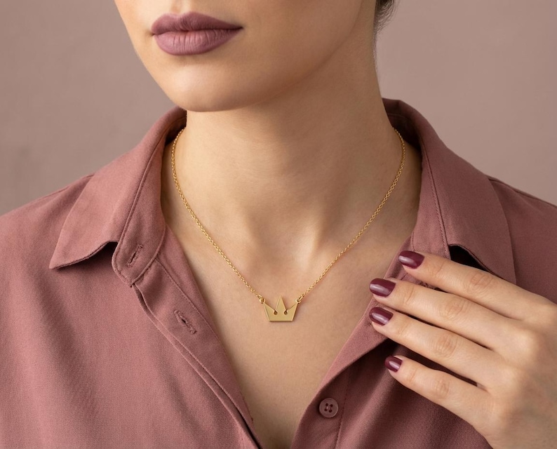 May include: A gold-colored crown pendant necklace. The necklace features a delicate chain and a small, stylized crown pendant. The person is wearing a pink collared shirt. The background is a solid, neutral color.