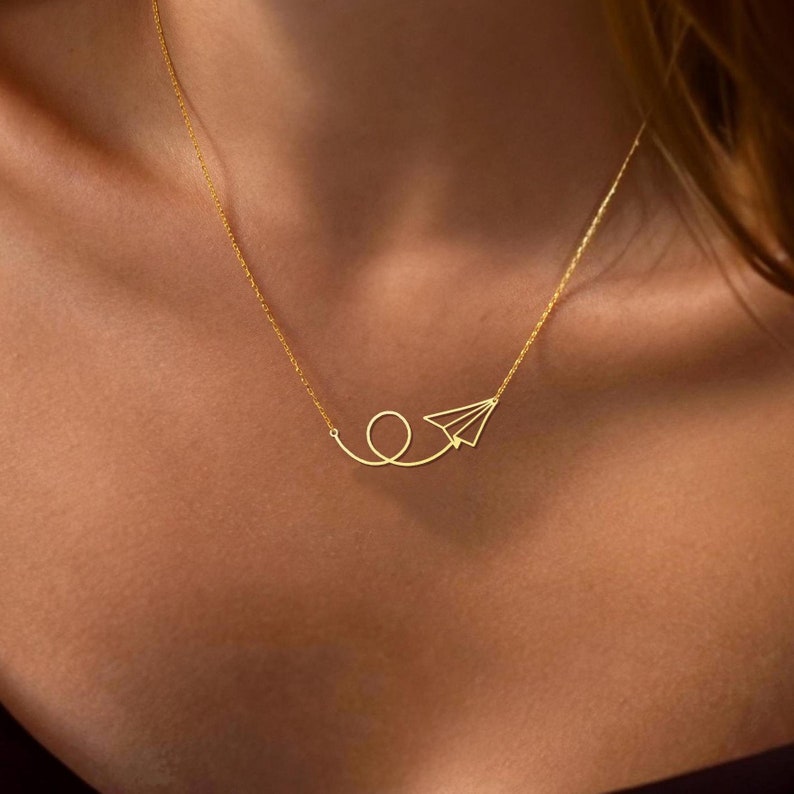 May include: A gold necklace with a delicate paper airplane pendant. The airplane is flying in a loop, symbolizing travel and freedom.