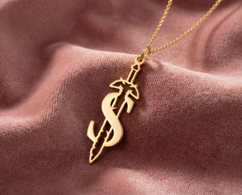 May include: A gold-colored pendant necklace featuring a stylized sword design with an "S" shape. The pendant hangs from a delicate gold chain. The necklace is displayed on a pinkish-brown velvet surface.