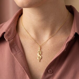 May include: A gold-colored pendant necklace featuring a dollar sign design with a sword through it. The pendant hangs from a delicate gold chain. The person in the image is wearing a pink button-down shirt.