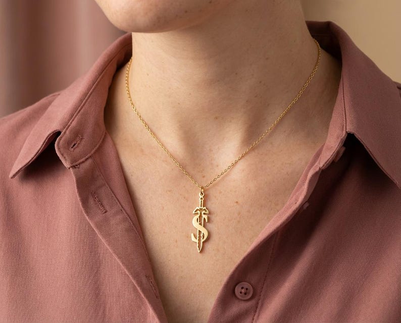 May include: A gold-colored pendant necklace featuring a dollar sign design with a sword through it. The pendant hangs from a delicate gold chain. The person in the image is wearing a pink button-down shirt.