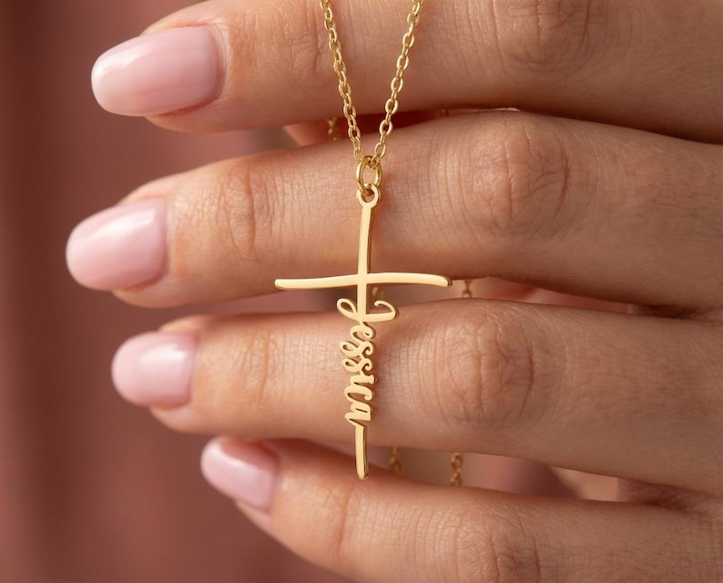 May include: A gold-coloured cross pendant necklace with the name "Jessica" inscribed vertically. The cross has a horizontal bar and a vertical bar, and the name is in a cursive font.
