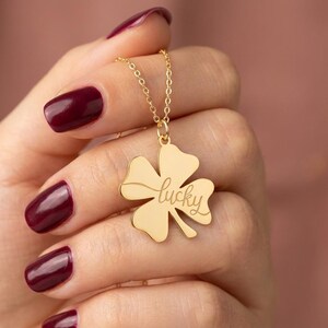 Pode incluir: Um colar dourado com um pingente de trevo de quatro folhas. O pingente tem a palavra "lucky" gravada. O colar &eacute; segurado contra um fundo desfocado. A pessoa que segura o colar tem esmalte de unha bord&ocirc;.