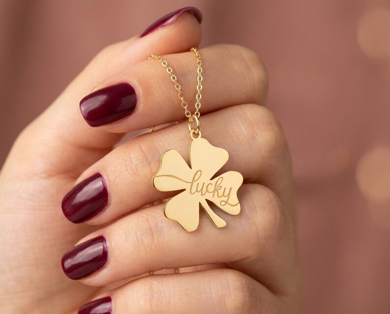 Pode incluir: Um colar dourado com um pingente de trevo de quatro folhas. O pingente tem a palavra "lucky" gravada. O colar &eacute; segurado contra um fundo desfocado. A pessoa que segura o colar tem esmalte de unha bord&ocirc;.