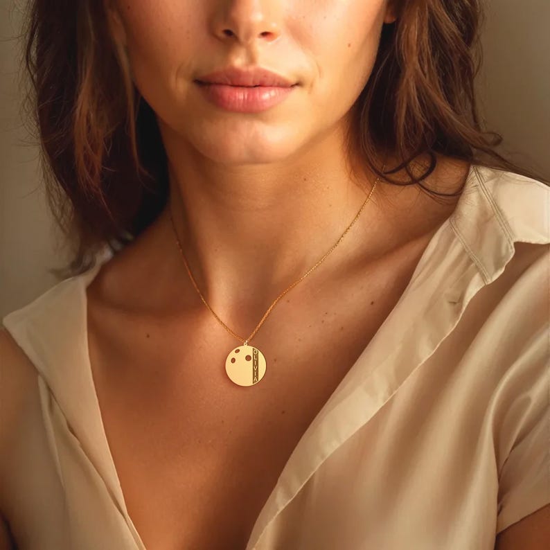 May include: A gold pendant necklace featuring a circular pendant. The pendant has a design with three dots and a vertical stripe. The necklace is worn around a person's neck, with a fine chain.