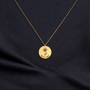 May include: A gold-coloured necklace with a circular pendant. The pendant has an engraved flower and the date 05.07.1997. The chain is delicate and hangs against a dark blue fabric background.