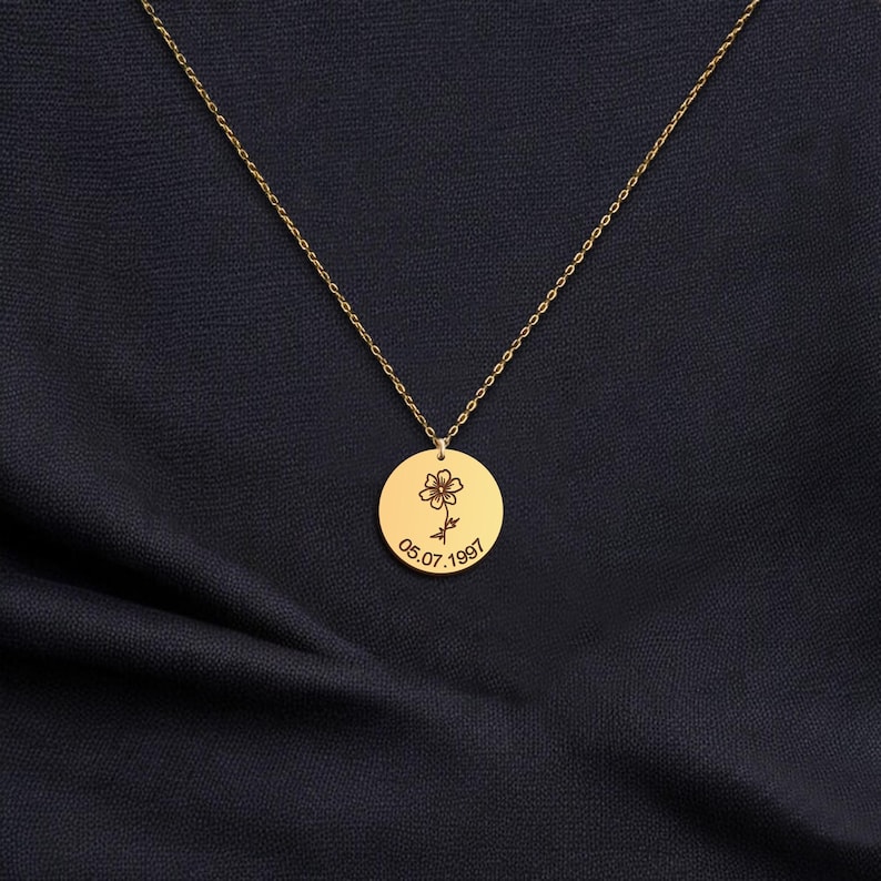 May include: A gold-coloured necklace with a circular pendant. The pendant has an engraved flower and the date 05.07.1997. The chain is delicate and hangs against a dark blue fabric background.