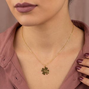 May include: A gold-colored necklace with a four-leaf clover pendant. The pendant has the word "lucky" engraved on it. The necklace is worn by a person wearing a pink shirt.