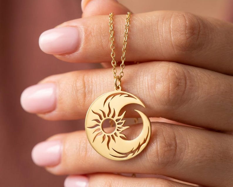 Op de afbeelding: Een goudkleurige ketting met een hanger met een zon- en maanontwerp. De ronde hanger heeft een zonnepatroon in het midden en een halve maanvorm aan de zijkant. De ketting wordt door een persoon vastgehouden.