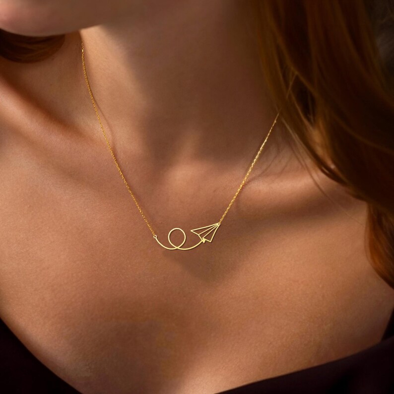 May include: A gold necklace with a paper airplane pendant. The airplane is flying in a loop.
