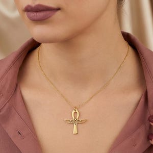 May include: A gold-colored Ankh pendant necklace. The pendant features a cross-like shape with a loop at the top and stylized wings. The necklace is worn on a person with a pink shirt.