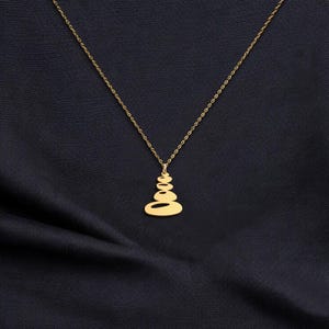 K&ouml;nnte beinhalten: Eine goldfarbene Halskette mit Anh&auml;nger, die ein Design aus gestapelten Steinen aufweist. Der Anh&auml;nger h&auml;ngt an einer feinen Kette. Die Halskette wird vor einem dunkelblauen Stoffhintergrund pr&auml;sentiert, der einen Kontrast erzeugt.