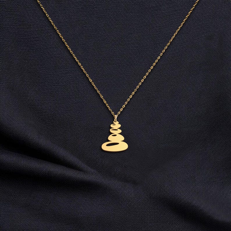 K&ouml;nnte beinhalten: Eine goldfarbene Halskette mit Anh&auml;nger, die ein Design aus gestapelten Steinen aufweist. Der Anh&auml;nger h&auml;ngt an einer feinen Kette. Die Halskette wird vor einem dunkelblauen Stoffhintergrund pr&auml;sentiert, der einen Kontrast erzeugt.