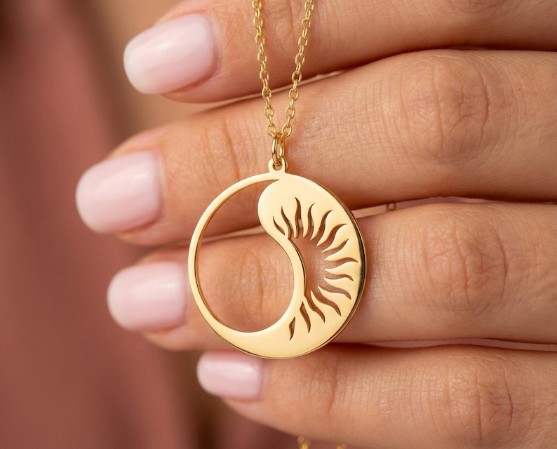 May include: A gold-colored pendant necklace featuring a sun and moon design. The pendant has a circular shape with a sunburst pattern on one side and a crescent moon shape on the other. The necklace chain is also gold.
