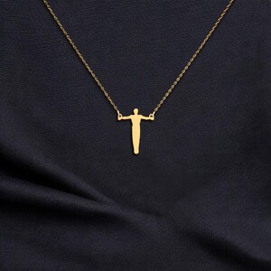 May include: A gold pendant necklace with a stylized human figure design. The pendant, with arms outstretched, hangs from a fine chain. The necklace is set against a dark blue fabric backdrop.