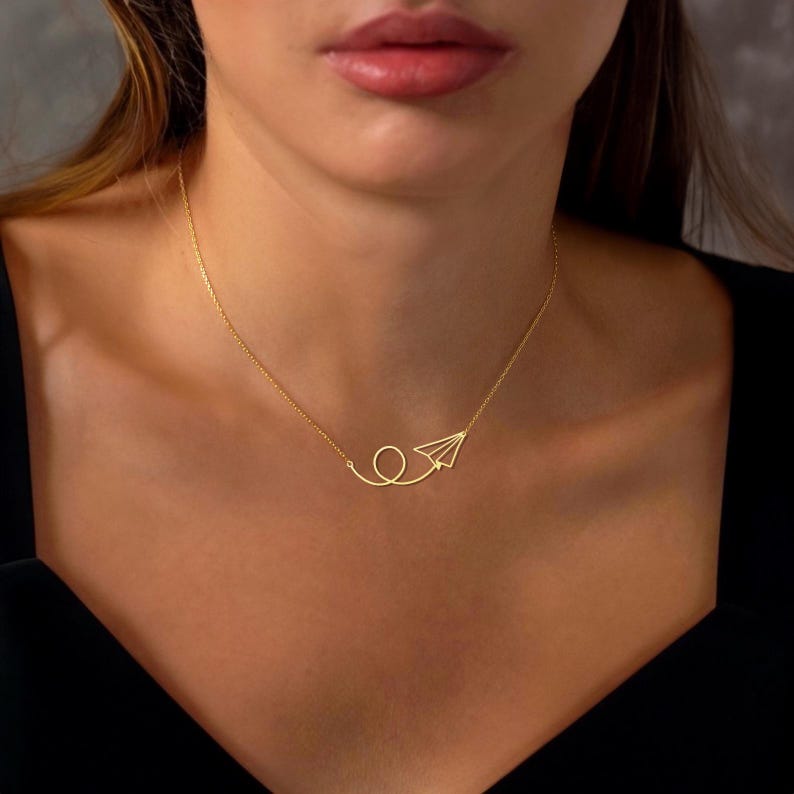 May include: A gold necklace with a paper airplane pendant. The airplane is made of thin metal and is flying in a loop.
