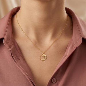May include: A gold-colored necklace with a pendant featuring a sun design within a crescent moon shape. The necklace is worn against a pink button-down shirt.