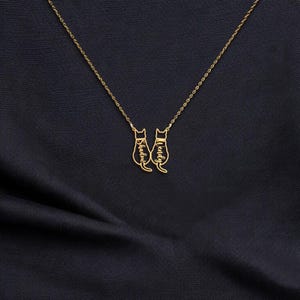 May include: A gold-colored necklace featuring two cat pendants. Each cat pendant has a name engraved on it. The necklace is displayed on a dark blue fabric background.