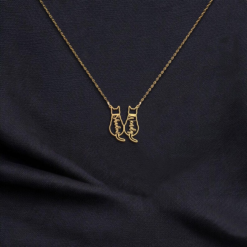 May include: A gold-colored necklace featuring two cat pendants. Each cat pendant has a name engraved on it. The necklace is displayed on a dark blue fabric background.