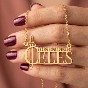 May include: Gold-colored necklace with the text "THE LEGEND OF CELES" in a stylized font. The pendant features a sword design integrated into the letter "C". The necklace is held by a hand with dark red nail polish.