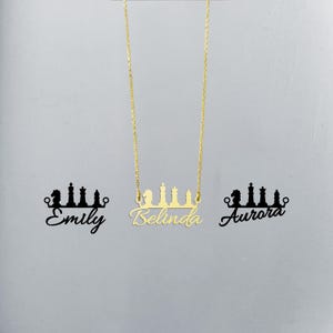 May include: Gold necklace featuring a chess piece design and the name "Belinda" written in cursive. The necklace is displayed against a white background with the names "Emily" and "Aurora" written in black below the necklace.