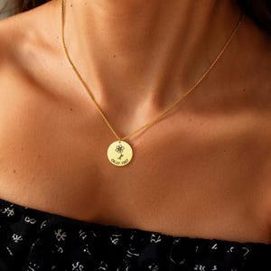 May include: A gold necklace with a circular pendant. The pendant has a small flower design and the date "05.07.1997" engraved. The necklace is worn on a person's chest. The chain and pendant are gold in colour.