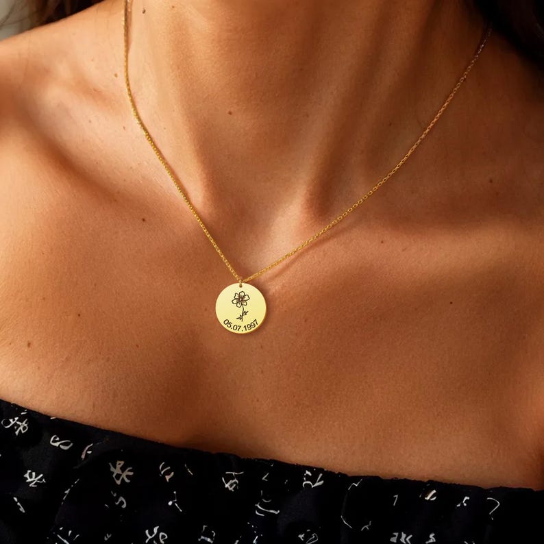 May include: A gold necklace with a circular pendant. The pendant has a small flower design and the date "05.07.1997" engraved. The necklace is worn on a person's chest. The chain and pendant are gold in colour.