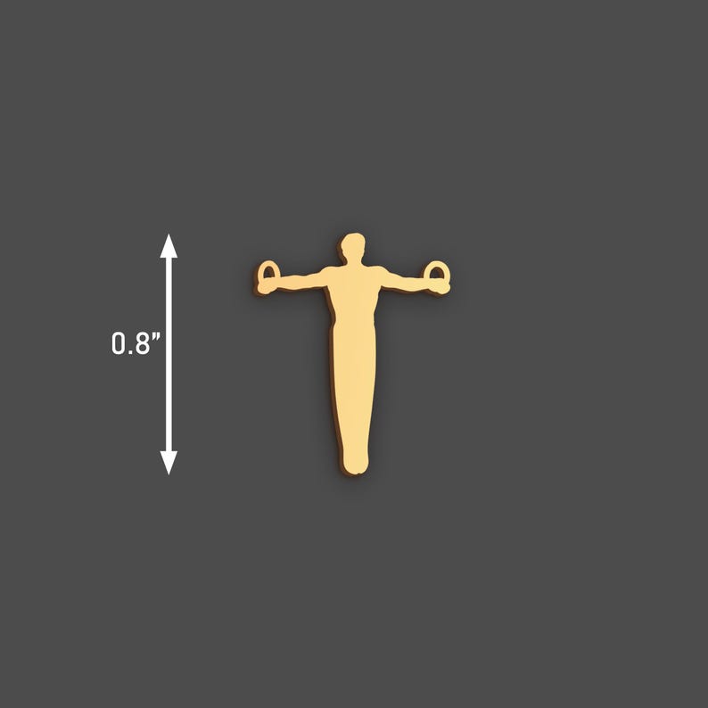 May include: A gold-coloured gymnast figure with arms outstretched, holding rings. The figure is approximately 2 cm tall. The background is a solid dark grey. This item is likely a charm or pendant.