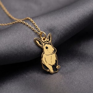May include: A gold-toned metal necklace featuring a pendant shaped like a rabbit. The rabbit pendant has black enamel detailing on the ears and lower body, with a smooth gold finish on the face and chest. It hangs from a delicate gold-toned chain.