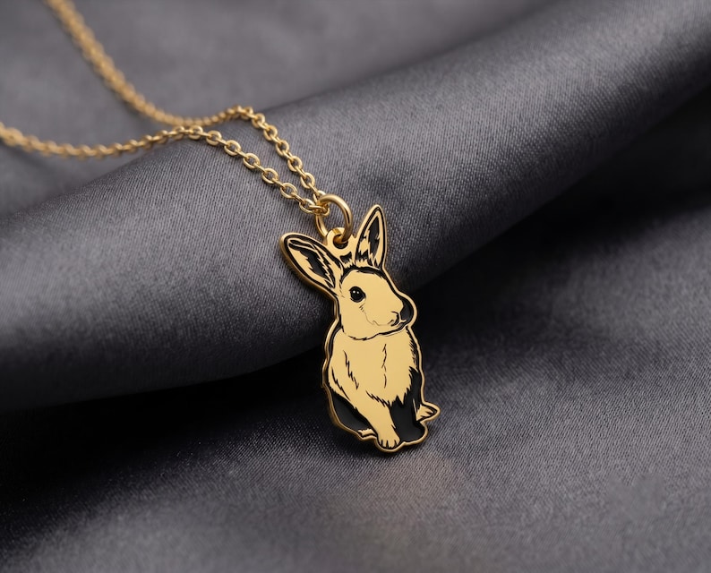 May include: A gold-toned metal necklace featuring a pendant shaped like a rabbit. The rabbit pendant has black enamel detailing on the ears and lower body, with a smooth gold finish on the face and chest. It hangs from a delicate gold-toned chain.