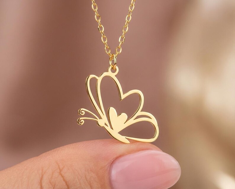 May include: A gold-colored butterfly pendant necklace. The butterfly design is open, with a heart-shaped center. The necklace chain is also gold and delicate. The pendant is held by a person with a pink nail polish.