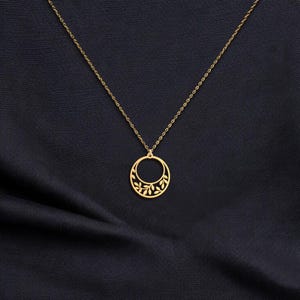 Puede incluir: Un collar dorado con un delicado colgante circular. El colgante presenta un dise&ntilde;o recortado de hojas y est&aacute; suspendido de una fina cadena. El collar se muestra sobre un fondo de tela azul oscuro.