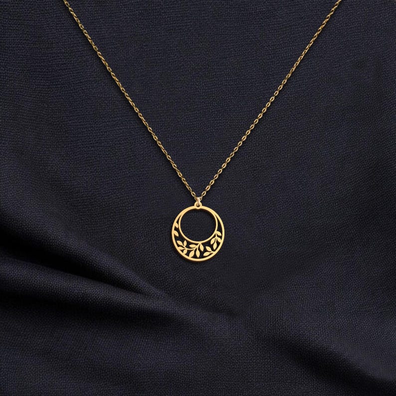 Puede incluir: Un collar dorado con un delicado colgante circular. El colgante presenta un dise&ntilde;o recortado de hojas y est&aacute; suspendido de una fina cadena. El collar se muestra sobre un fondo de tela azul oscuro.