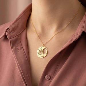 Puede incluir: Un collar con colgante dorado con un dise&ntilde;o de sol y luna creciente. El colgante circular presenta un patr&oacute;n de sol y una forma de luna curvada. El collar se lleva sobre una camisa rosa abotonada.