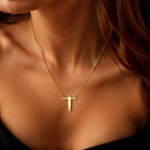 May include: A gold necklace with a pendant in the shape of a person with arms outstretched. The pendant is suspended from a fine chain. The necklace is worn against skin, with a dark background.
