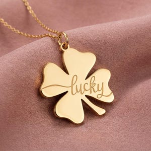 May include: A gold-colored necklace with a four-leaf clover pendant. The pendant has the word "lucky" engraved in a cursive font. The necklace is displayed on a pink fabric background.