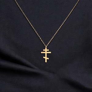 May include: A gold-colored Orthodox cross pendant necklace. The cross features a three-bar design, suspended from a delicate chain. The necklace is displayed against a dark, textured background, highlighting the pendant's details.