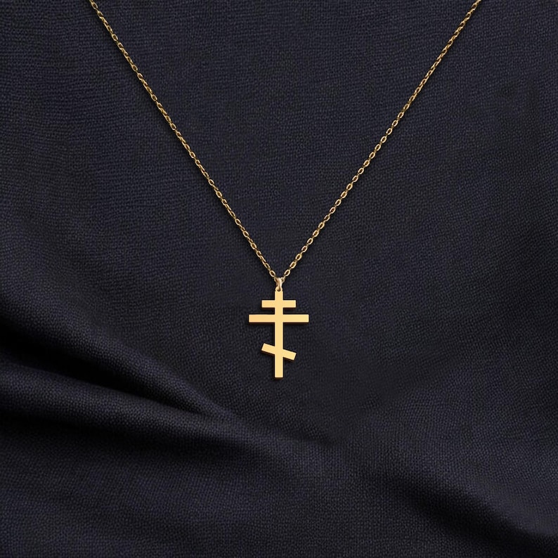 May include: A gold-colored Orthodox cross pendant necklace. The cross features a three-bar design, suspended from a delicate chain. The necklace is displayed against a dark, textured background, highlighting the pendant's details.