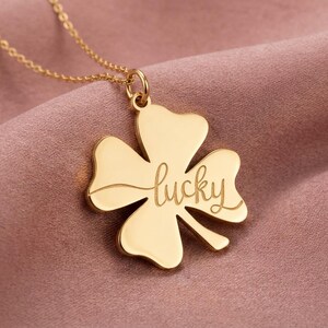 Pode incluir: Um colar dourado com um pingente de trevo de quatro folhas. O pingente tem a palavra "lucky" gravada em it&aacute;lico. O colar &eacute; exibido em um fundo de tecido rosa.