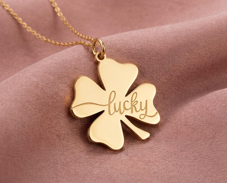 Pode incluir: Um colar dourado com um pingente de trevo de quatro folhas. O pingente tem a palavra "lucky" gravada em it&aacute;lico. O colar &eacute; exibido em um fundo de tecido rosa.