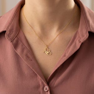 May include: A gold-colored butterfly pendant necklace. The butterfly design is delicate and detailed, with a small gemstone accent. The necklace is worn against a pink button-down shirt.