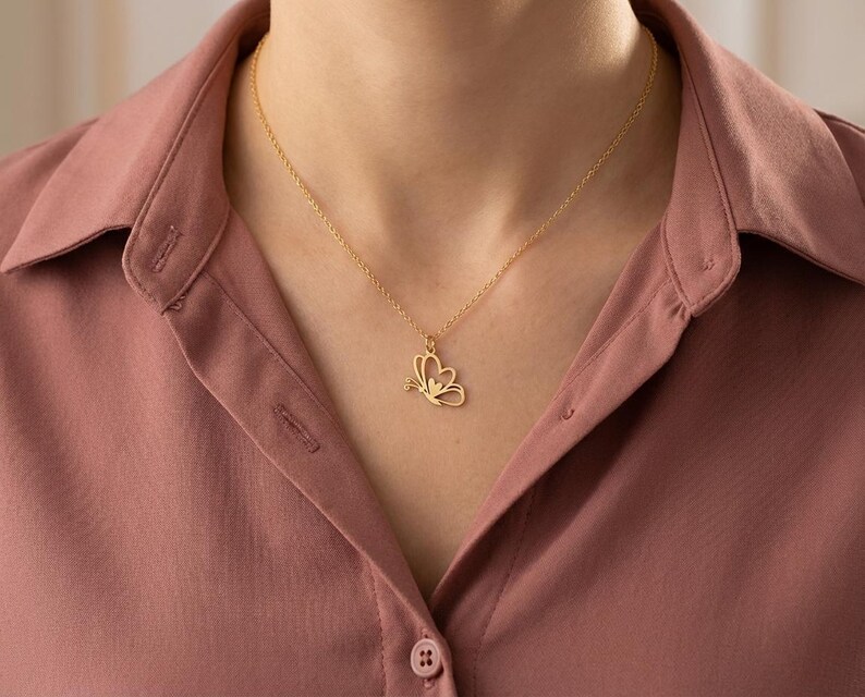 May include: A gold-colored butterfly pendant necklace. The butterfly design is delicate and detailed, with a small gemstone accent. The necklace is worn against a pink button-down shirt.